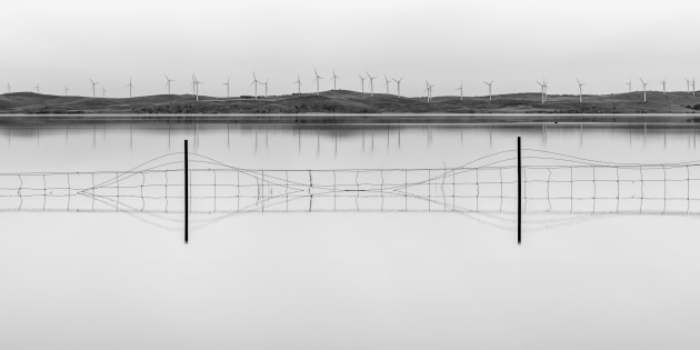 Places category winner, Wade Cramer, Flooded Fences. Wade tells us his striking minimalist image Flooded Fences was taken last year at Lake George on the way to Canberra. “We stopped as we had never seen the lake so calm,” he says. 
“There was such great light, and when I took the shot I had a monochrome image in mind. There is a beauty to the fence wires as they flow in and out of the water and then there is that juxtaposition between the flooding caused by lots of rain, a symptom of climate change, and the wind turbines as is a possible solution to climate change,” he says.